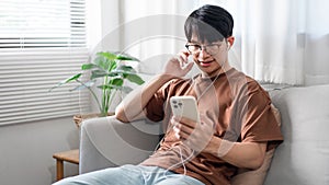 Technology Concept The person who wears glasses and headphones sitting on the grey couch and listening music on his smartphone