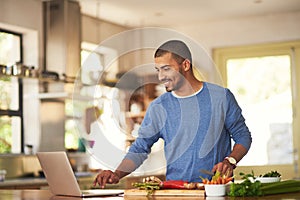 Technology has its place in the kitchen. Shot of a happy young man using a laptop while preparing a healthy meal at home