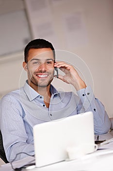 Technology keeps him at the top of his game. a smiling young man sitting at his desk and using his mobile phone.