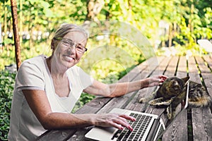 Happy senior old woman with home cat working online with laptop computer outdoor