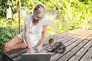 Happy senior old woman with home cat working online with laptop computer outdoor
