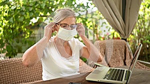 Technology, old age people concept - elderly happy senior woman puts on protective face mask working online with laptop computer