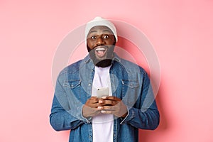 Technology and online shopping concept. Surprised young Black man using mobile phone, looking at camera amazed and happy