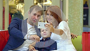 Technology and people concept - happy young couple with shopping bags and smartphone taking selfie in mall. Lovely