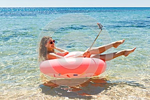 Technology and people concept - smiling young woman or teenage girl makes selfie on inflatable donut with smartphone on beach