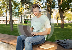 Technology and studentship concept. Male student using laptop computer and wearing headphones, sitting outdoors