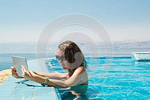 Technology and vacation concept. Luxury travel. Young pretty woman using tablet computer in infinity pool at resort.
