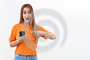 Technology, youth and communication concept. Portrait of excited, surprised blond girl pointing finger mobile phone