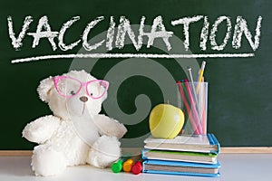 Teddy bear next to a pile of books and an apple in a classroom. Blackboard for the vaccination period concept