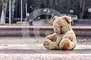 A teddy bear in the park sits on an idle fountain, a woman with a stroller leaves along the park alley.