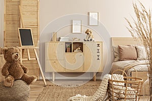Teddy bear on pouf and small blackboard on easel in stylish kid`s bedroom with wooden cabinet and beige bedding on bed