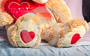 A teddy bear is sitting on the bed with hearts on its feet holding another heart that says `Be my valentine`. Close up