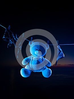 A teddy bear toy hangs on a clothesline on clothespins in neon light. tied with shibari ropes