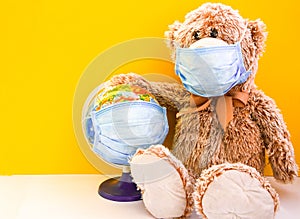 Teddy bear toy wearing medical face mask and holding earth world globe with protective mask on yellow background, copy