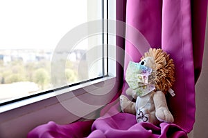 A teddy bear waering a medical protective mask sits on a windowsill and looks out the window. Sadness and loneliness during