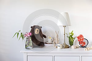 A teddy bear, a wooden hare,  bouquet of flowers, books on a white chest of drawers in the interior of the room