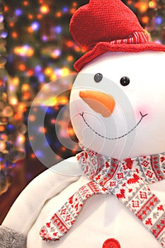 Teddy puppet snowman with red hat, scarf and orange carrot nose. Blurred colored lights background