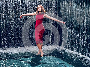 Teen ager girl in high fashion red dress posing in water, water fall behind her back. Prom photoshoot in beautiful location Young