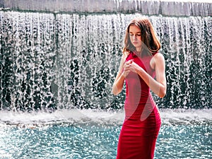 Teen ager girl in high fashion red dress posing in water, water fall behind her back. Prom photoshoot in beautiful location Young