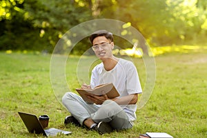 Teen Asian Guy Studying With Book Outdoors In Park And Taking Notes