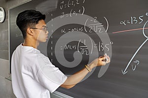 Teen asian high school boy student solving maths exercise on blackboard with chalk