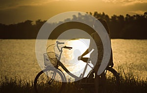 teen asian woman sit at retro vintage bicycle near the lake at sunset moment. silhouette bicycle at the sunset with grass field.