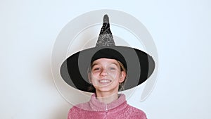 A teen blond girl in black witch hat is actively telling something and looking at the camera. The girl sings or recites