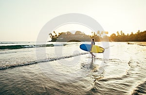 Teen boy with blue and yellow surfboard entering the waves for surfing with sunset rays on palm trees grove. Happy childhood and