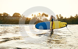 Teen boy with blue and yellow surfboard entering the waves for surfing with sunset rays on palm trees grove. Happy childhood and