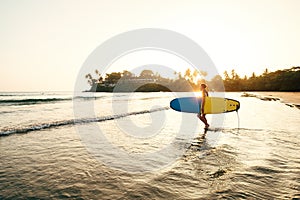 Teen boy with blue and yellow surfboard entering the waves for surfing with sunset rays on palm trees grove. Happy childhood and