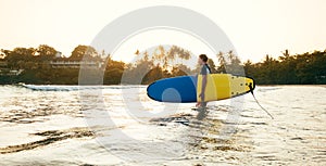 Teen boy with blue and yellow surfboard entering the waves for surfing with sunset rays on palm trees grove. Happy childhood and