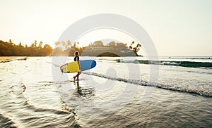 Teen boy with blue and yellow surfboard entering the waves for surfing with sunset rays on palm trees grove. Happy childhood and