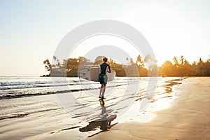 Teen boy with blue and yellow surfboard entering the waves for surfing with sunset rays on palm trees grove. Happy childhood and