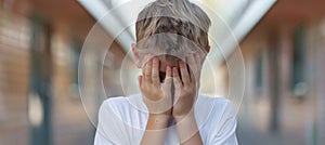 Teen boy in corridor, hands covering face, learning difficulties, blurred background, text space.