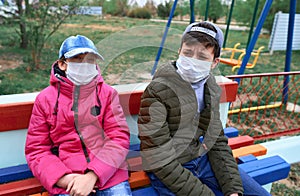 Teen boy and girl sitting on a bench on playground near high-rise buildings with apartments, a medical mask on their faces