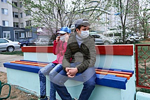 Teen boy and girl sitting on a bench on playground near high-rise buildings with apartments, a medical mask on their faces