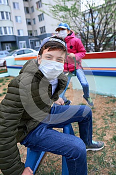 Teen boy and girl sitting on a seesaw on playground near high-rise buildings with apartments, a medical mask on their faces