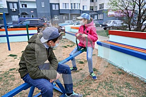 Teen boy and girl sitting on a seesaw on playground near high-rise buildings with apartments, a medical mask on their faces