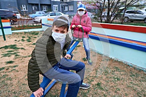 Teen boy and girl sitting on a seesaw on playground near high-rise buildings with apartments, a medical mask on their faces