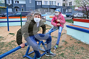 Teen boy and girl sitting on a seesaw on playground near high-rise buildings with apartments, a medical mask on their faces