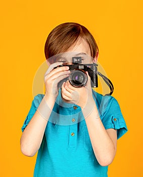 Teen boy looks through a vintage camera and trying the photographer profession. Studio portrait over yellow background