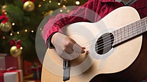 Teen boy playing guitar, sitting indoor near decorated xmas tree with lights, dressed as Santa helper - Merry Christmas and Happy