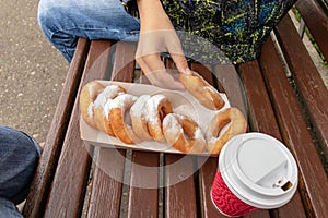 A teen boy sitting on a bench in park and eating beignet pastries, donuts and drinking coffee or tea from a takeaway box