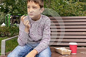 A teen boy sitting on a bench in park and eating beignet pastries, donuts and drinking coffee or tea from a takeaway box