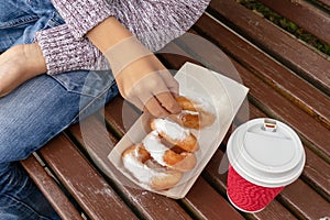 A teen boy sitting on a bench in park and eating beignet pastries, donuts and drinking coffee or tea from a takeaway box and cup,