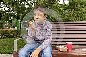 A teen boy sitting on a bench in park and eating beignet pastries, donuts and drinking coffee or tea from a takeaway box and cup,