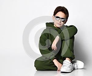 Teen boy in sportswear and sunglasses sitting on a gray background.