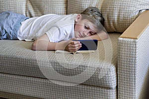Teen boy is using phone with open book lying on the sofa.