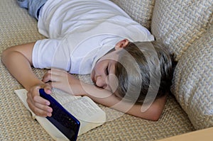 Teen boy is using phone with open book lying on the sofa.