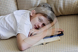 Teen boy is using phone with open book lying on the sofa.